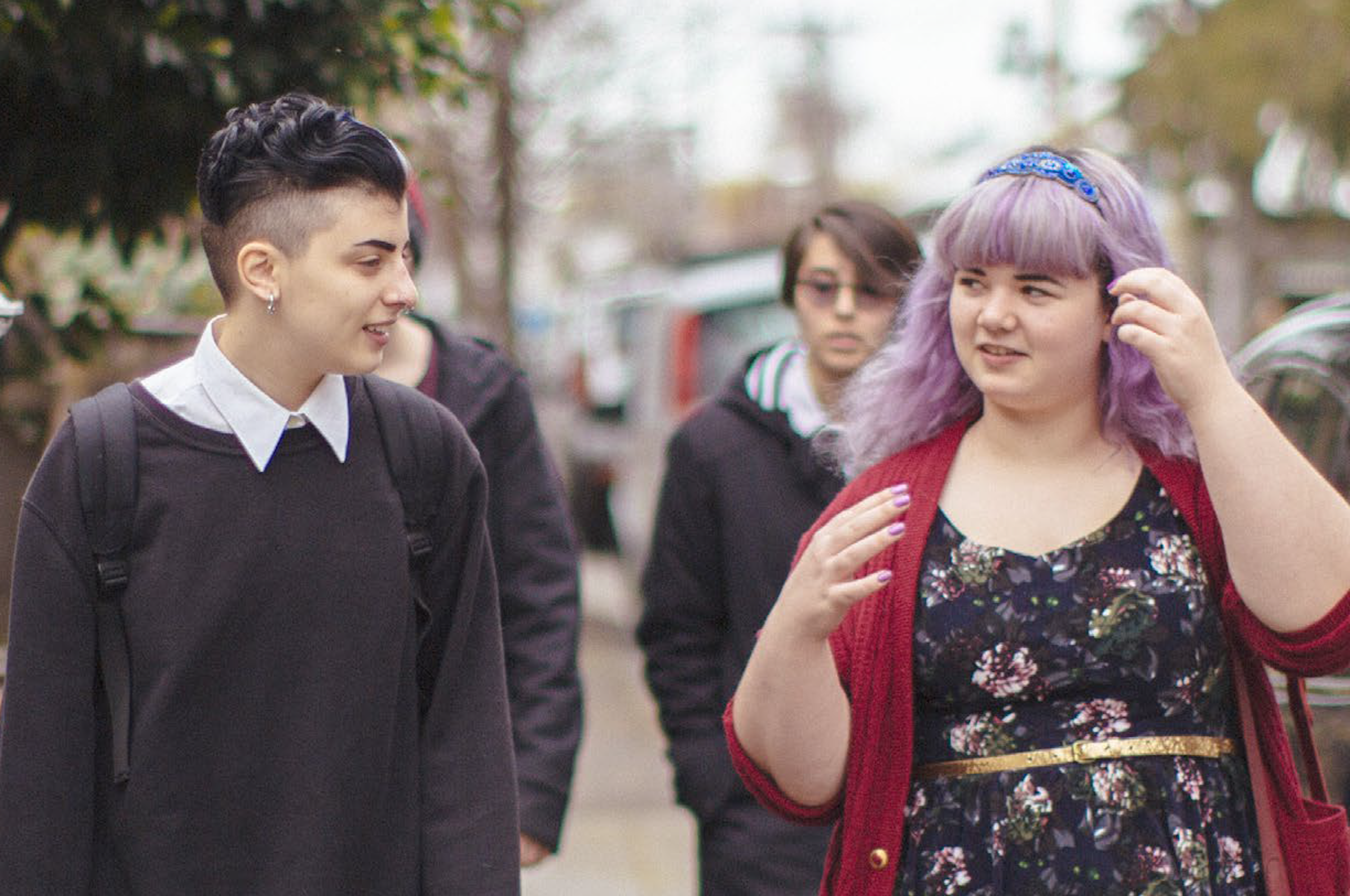 Two non-binary young people walking down a street, one with long purple hair and a floral dress and one with an undercut, shirt and jumper, earring and septum piercing