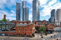 Photograph of a city skyline against a blue sky with puffy clouds, featuring several tall, sharp skyscrapers behind a group of heritage red-brick train station and bridge