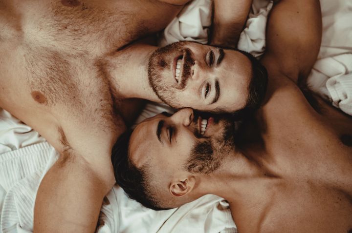 Two naked men lying in bed together, top and tailing, smiling and looking at the camera