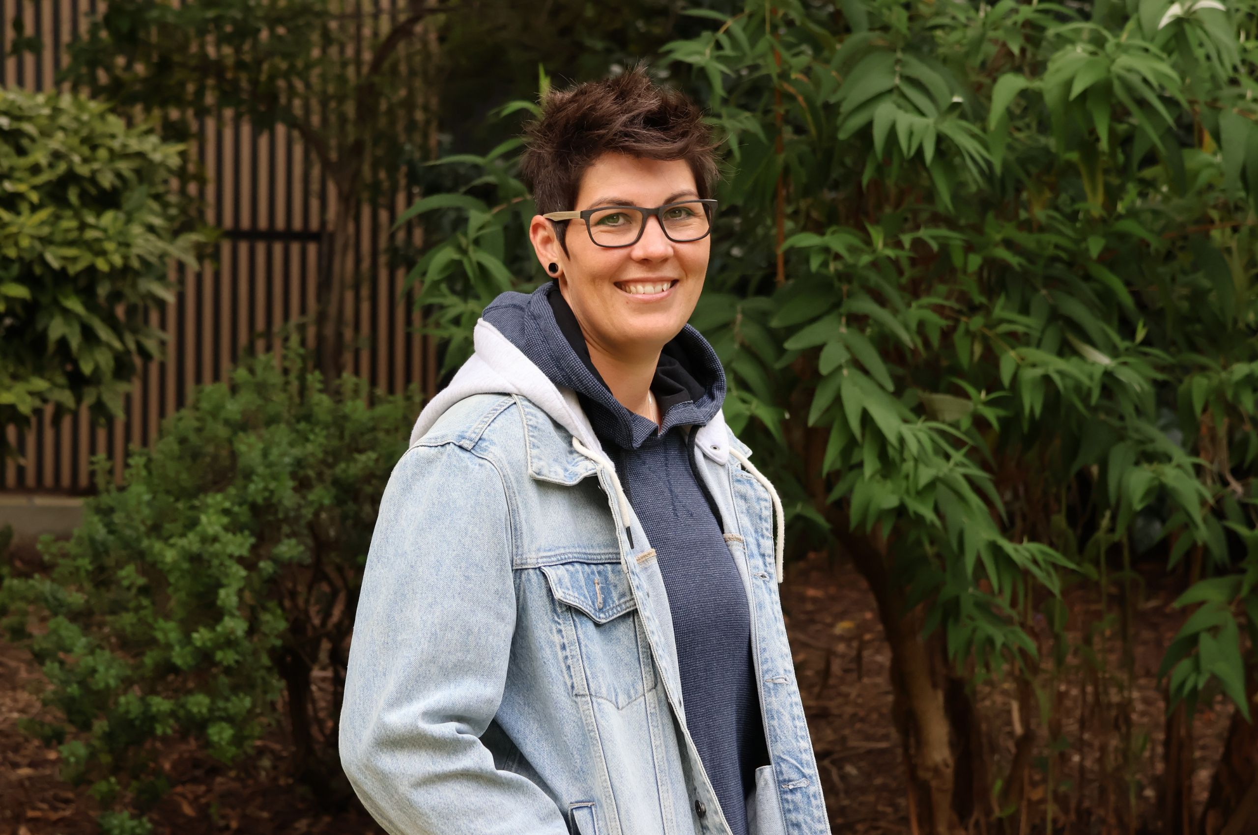 A woman with glasses wearing a denim jacket, smiling and looking confidently at the camera.