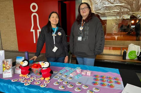 Two women standing behind a table of merchandise with a trans flag tablecloth