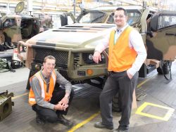 2 males in reflective vests smiling at camera standing in front of military camouflage vehicle